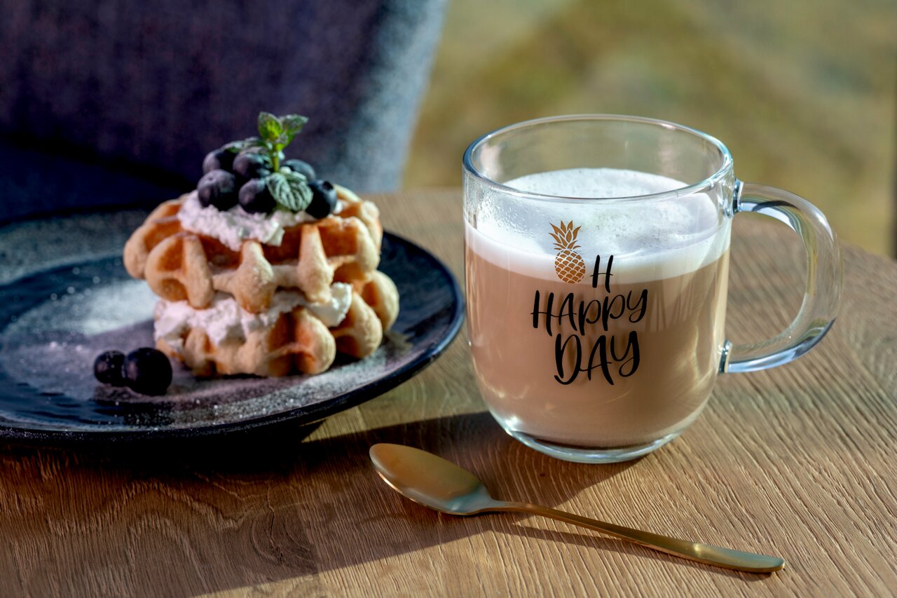Glazen beker met het opschrift 'Oh Happy Day' en een ananasmotief, gevuld met een romig drankje, op een houten tafel naast een bord met wafels en bessen, zijaanzicht.