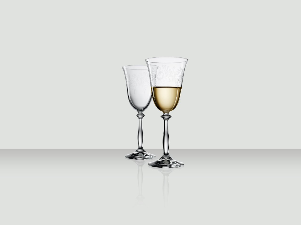 Zwei elegante Weingläser, eines gefüllt mit Weißwein, auf einer reflektierenden Oberfläche, seitliche Perspektive.