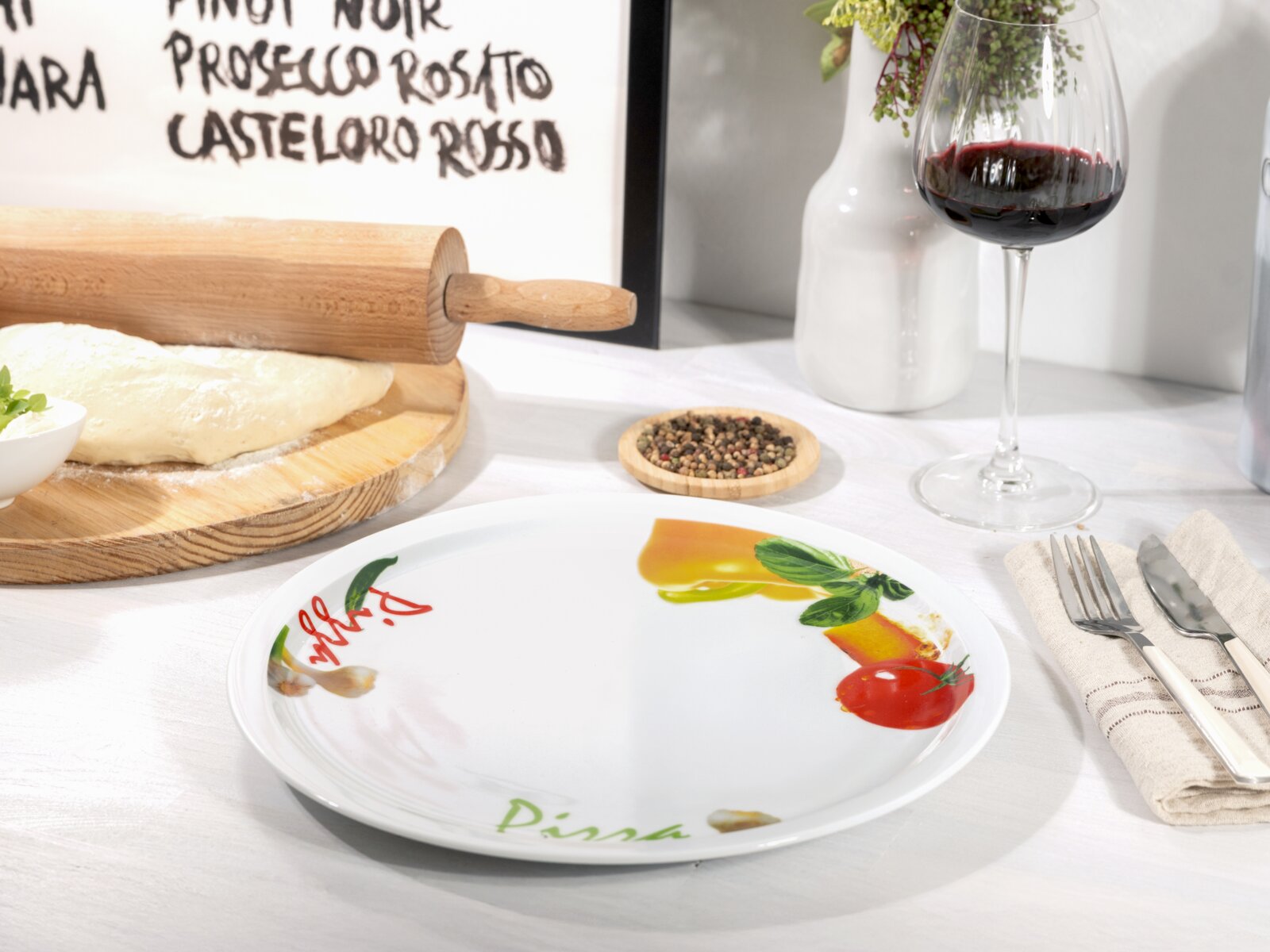 Runder weißer Pizzateller mit farbigem Pizza-Motiv und Schriftzug „Pizza“, in leicht schräger Vorderansicht auf einem gedeckten Tisch neben Besteck und Weinglas.