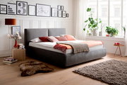 Modernes Polsterbett in Grau mit Kopfteil, dekoriert mit bunten Kissen und Decken, seitliche Perspektive im hellen Schlafzimmer mit Pflanzen und Wandbildern.