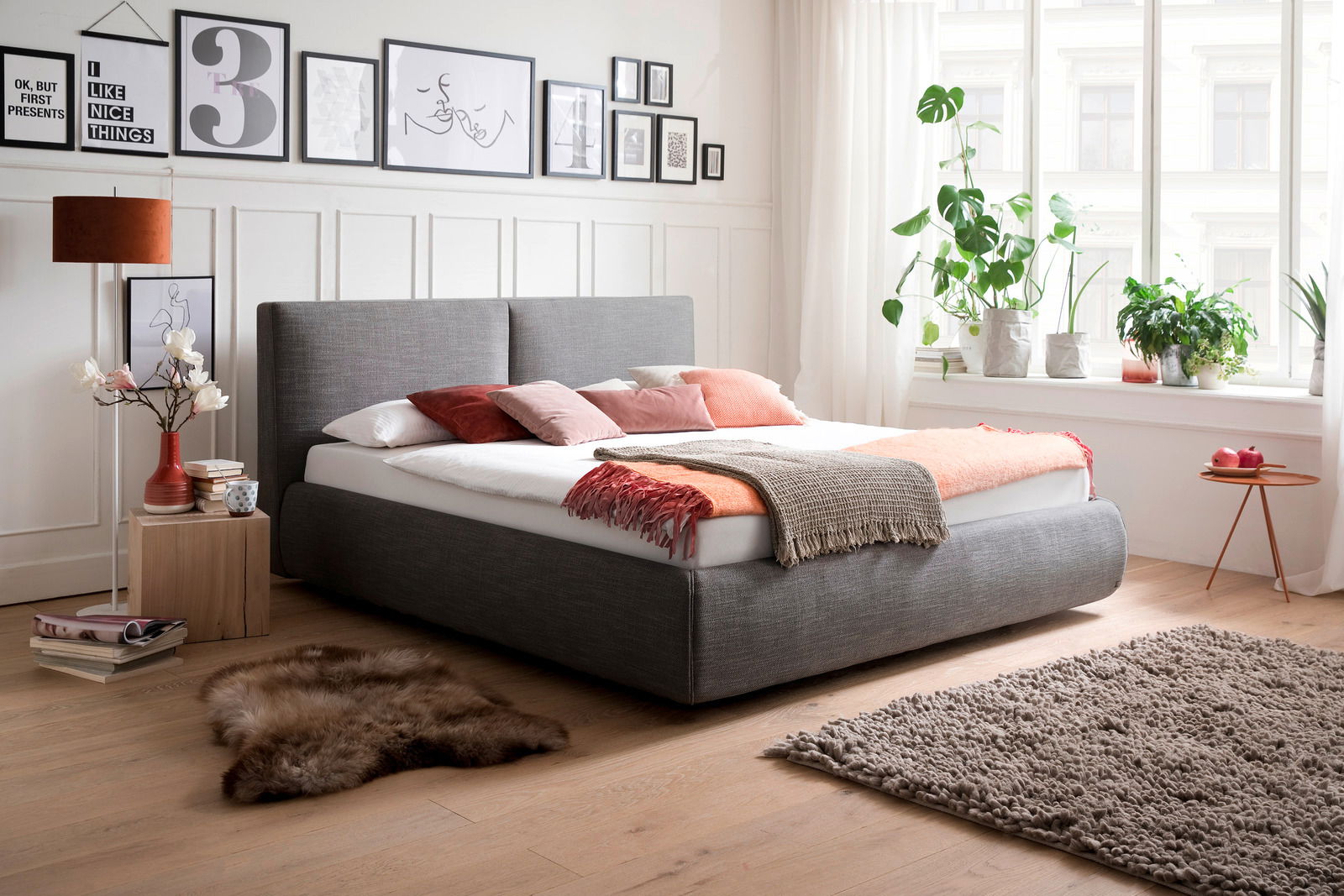Modernes Polsterbett in Grau mit Kopfteil, dekoriert mit bunten Kissen und Decken, seitliche Perspektive im hellen Schlafzimmer mit Pflanzen und Wandbildern.