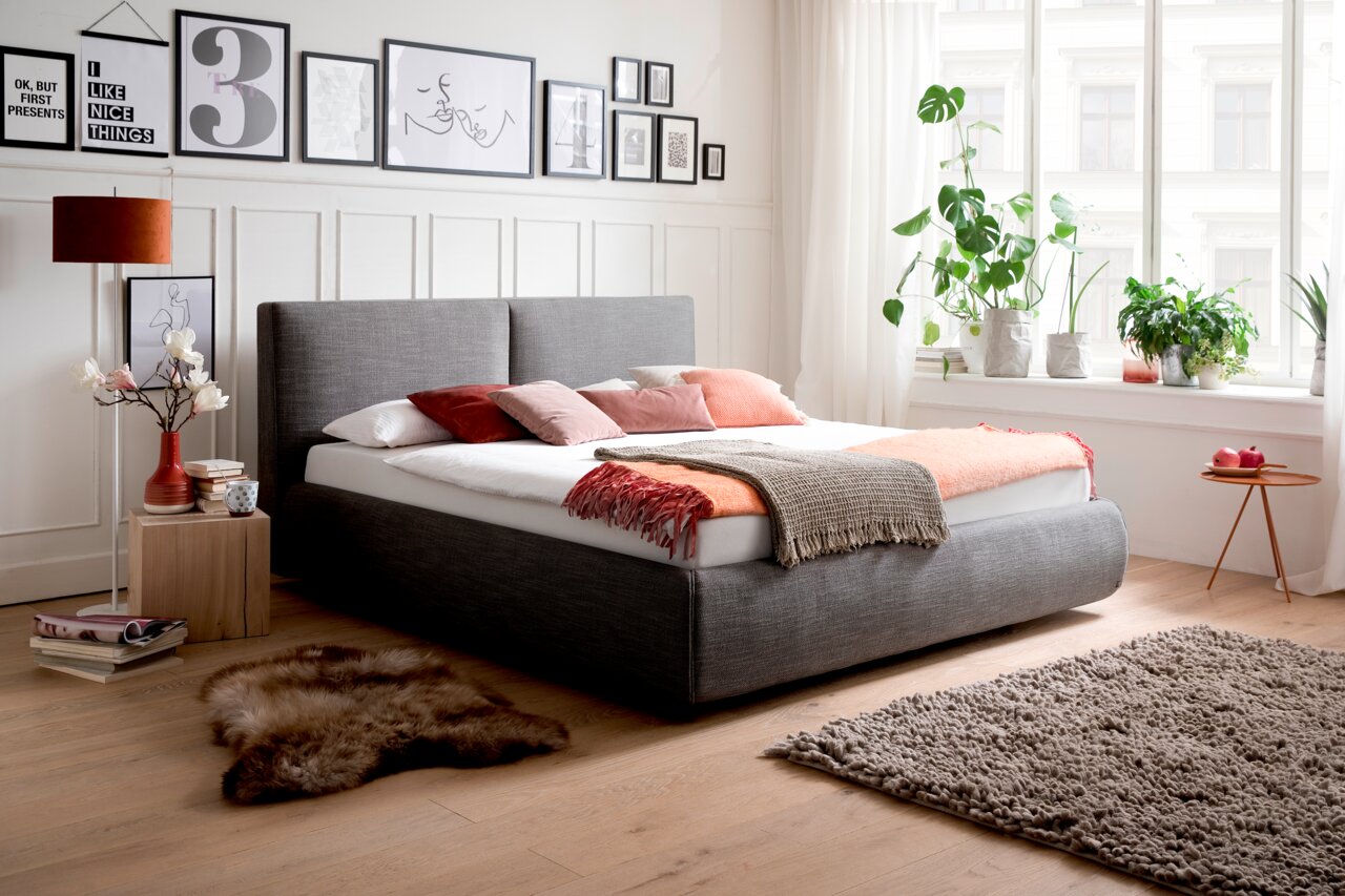 Modernes Polsterbett in Grau mit Kopfteil, dekoriert mit bunten Kissen und Decken, seitliche Perspektive im hellen Schlafzimmer mit Pflanzen und Wandbildern.