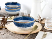 Blau-weiß glasierte Keramikschale mit braunem Rand auf hellem Holzbrett, gedeckter Tisch mit Besteck und Gläsern im Hintergrund, fotografiert in leicht schräger Vorderansicht