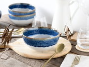 Blau-weiß glasierte Keramikschale mit braunem Rand auf hellem Holzbrett, gedeckter Tisch mit Besteck und Gläsern im Hintergrund, fotografiert in leicht schräger Vorderansicht