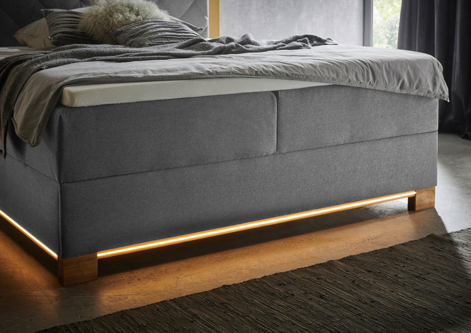 Boxspringbett in grauem Stoff mit Holzfüßen und integrierter Beleuchtung, seitliche Perspektive