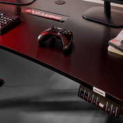 Gaming-Schreibtisch mit Carbon-Optik und BXGAMING-Logo, darauf ein schwarzer Gamecontroller; Ansicht schräg von oben (Top-Down-Perspektive) mit sichtbarem Bedienpanel an der Vorderkante.