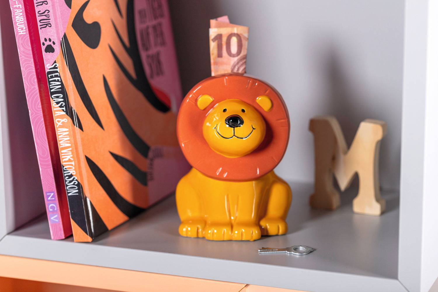 Spardose in Form eines sitzenden Löwen mit orangefarbener Mähne, frontal fotografiert, auf einem Regal platziert.