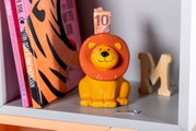 Spardose in Form eines sitzenden Löwen mit orangefarbener Mähne, frontal fotografiert, auf einem Regal platziert.