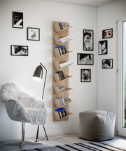 Schmales, wandmontiertes Bücherregal aus hellem Holz mit versetzt angeordneten Ablagen, befüllt mit Büchern und DVDs, in modernem Wohnzimmer – Frontansicht mit leicht seitlicher Perspektive.