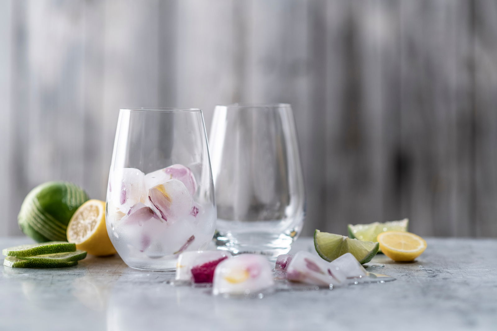 Zwei Universalbecher Forte, einer gefüllt mit Eiswürfeln und Zitrusfrüchten, auf einer Tischoberfläche mit geschnittenen Limetten und Zitronen im Vordergrund. Seitliche Perspektive.