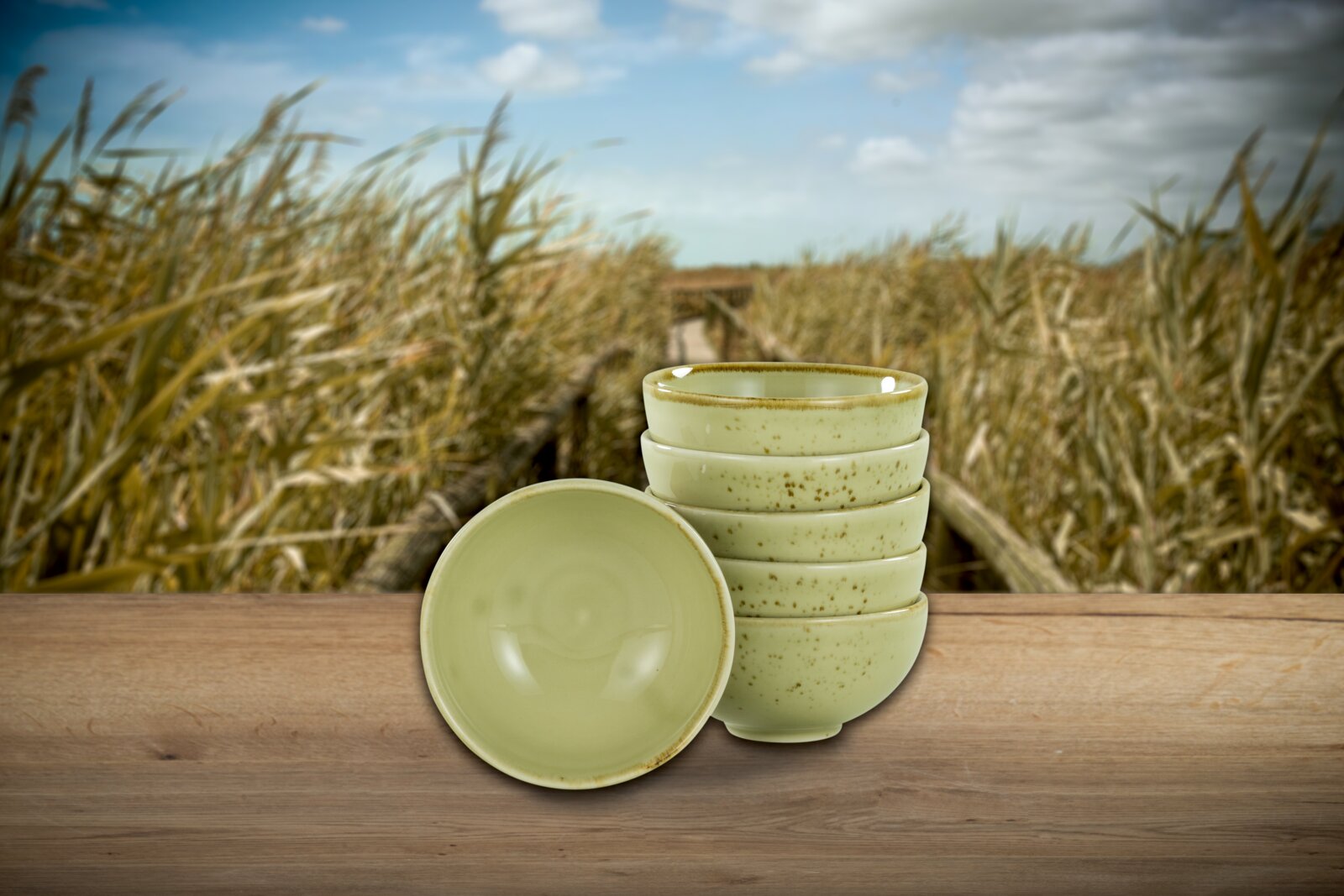 Stapel aus sechs hellgrünen, gesprenkelten Keramikschalen auf Holztisch, eine Schale vorne liegend Frontalansicht auf Augenhöhe vor Schilflandschaft und blauem Himmel