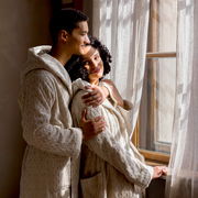 Zwei Personen in cremefarbenen Kapuzenmänteln stehen nebeneinander und schauen aus einem Fenster. Die Mäntel haben ein gestricktes Muster und wirken gemütlich. Die Szene ist von der Seite aufgenommen.