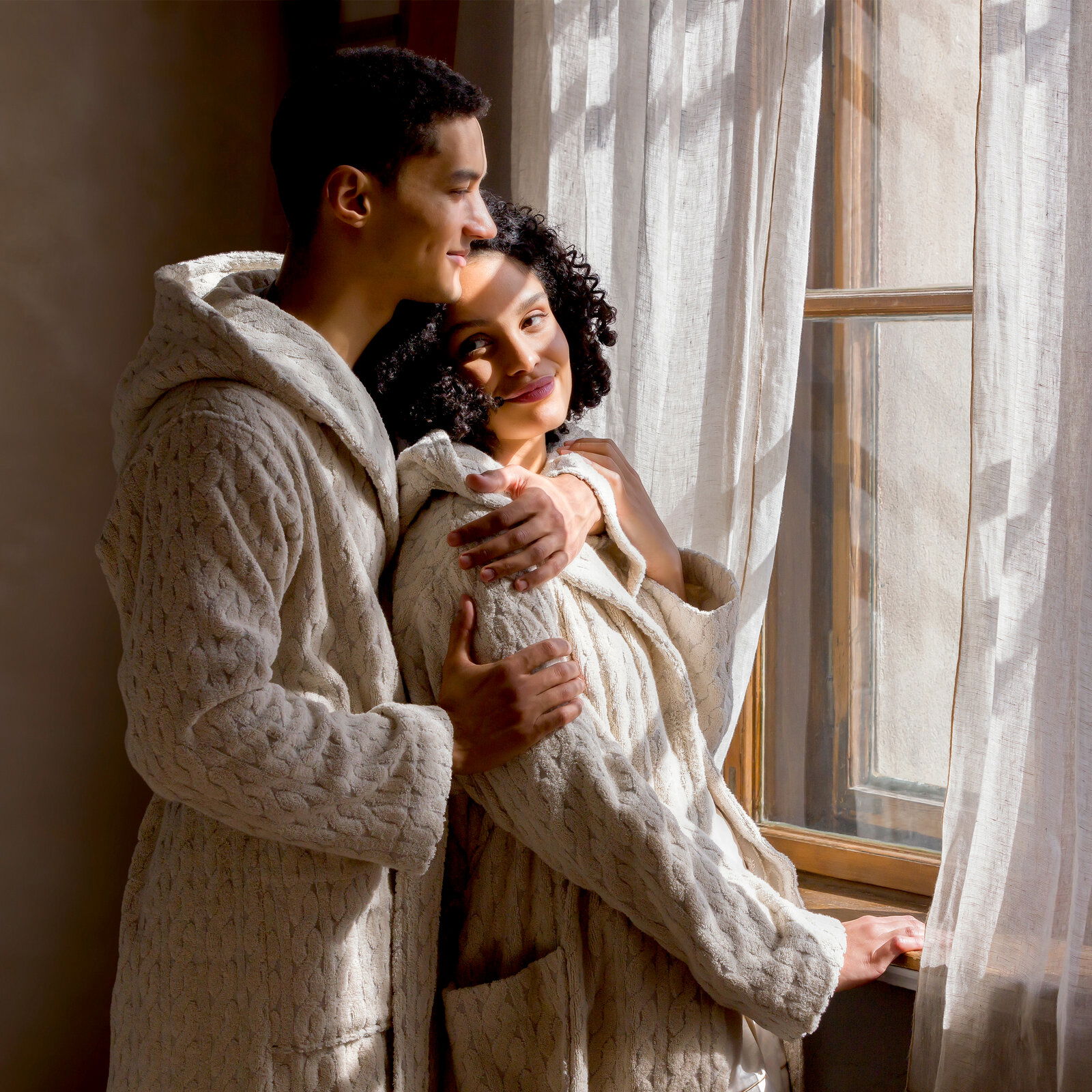 Zwei Personen in cremefarbenen Kapuzenmänteln stehen nebeneinander und schauen aus einem Fenster. Die Mäntel haben ein gestricktes Muster und wirken gemütlich. Die Szene ist von der Seite aufgenommen.