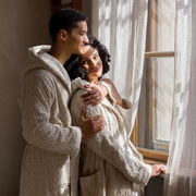 Zwei Personen in cremefarbenen Kapuzenmänteln stehen nebeneinander und schauen aus einem Fenster. Die Mäntel haben ein gestricktes Muster und wirken gemütlich. Die Szene ist von der Seite aufgenommen.