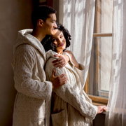 Zwei Personen in cremefarbenen Kapuzenmänteln stehen nebeneinander und schauen aus einem Fenster. Die Mäntel haben ein gestricktes Muster und wirken gemütlich. Die Szene ist von der Seite aufgenommen.