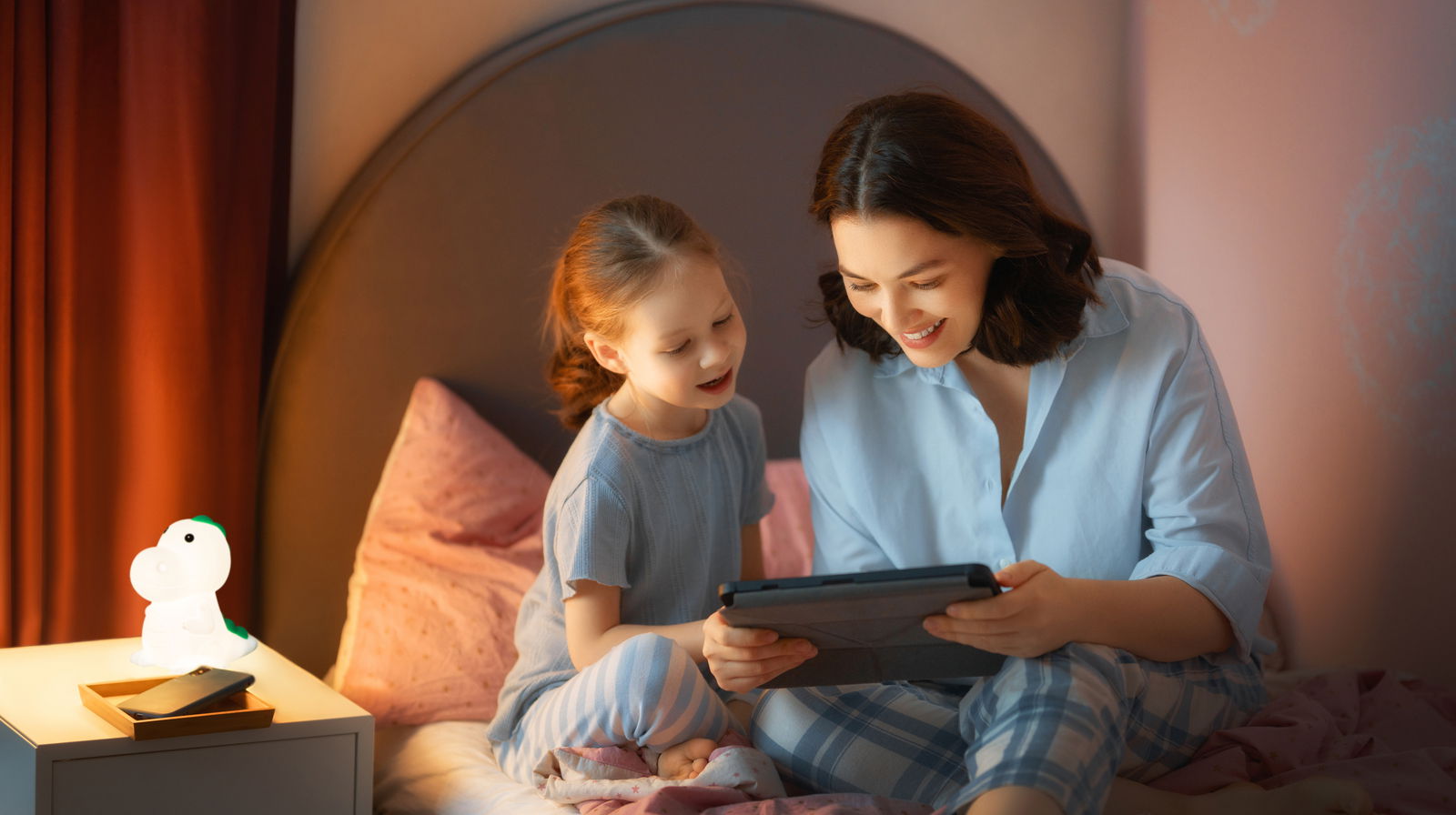 Eine Akku-Nachtleuchte in Form eines Dinosauriers steht auf einem Nachttisch neben einem Smartphone. Im Hintergrund sitzen eine Frau und ein Kind auf einem Bett und schauen gemeinsam auf ein Tablet. Die Szene ist in einem gemütlichen Schlafzimmer bei gedä