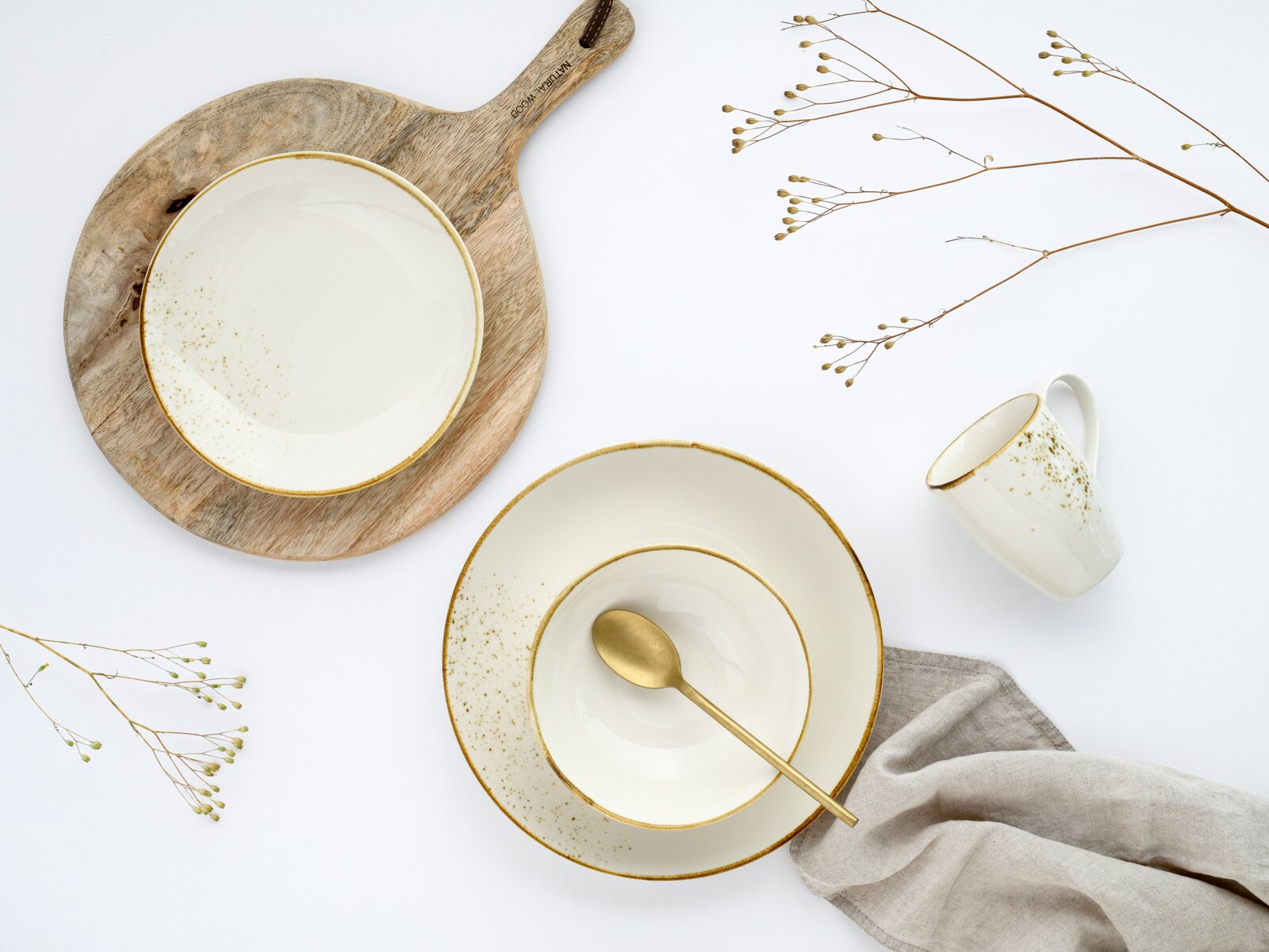 Weißes Keramik-Geschirrset mit goldfarbenem Rand und feinen Sprenkeln, bestehend aus Teller und Schale mit goldfarbenem Löffel sowie passender Tasse, arrangiert auf Holzbrett mit Leinentuch und Trockenblumen vor weißem Hintergrund, Draufsicht (Flatlay).