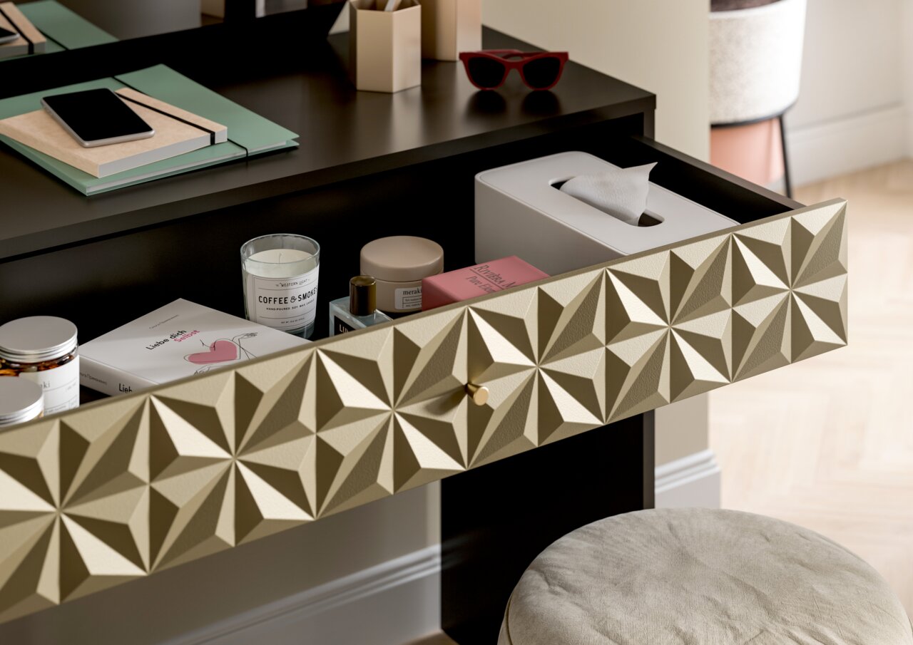 Nahaufnahme eines Schminktisches mit geöffneter Schublade, die verschiedene Kosmetikartikel und Accessoires enthält. Die Schubladenfront hat ein geometrisches Muster. Perspektive von schräg oben.
