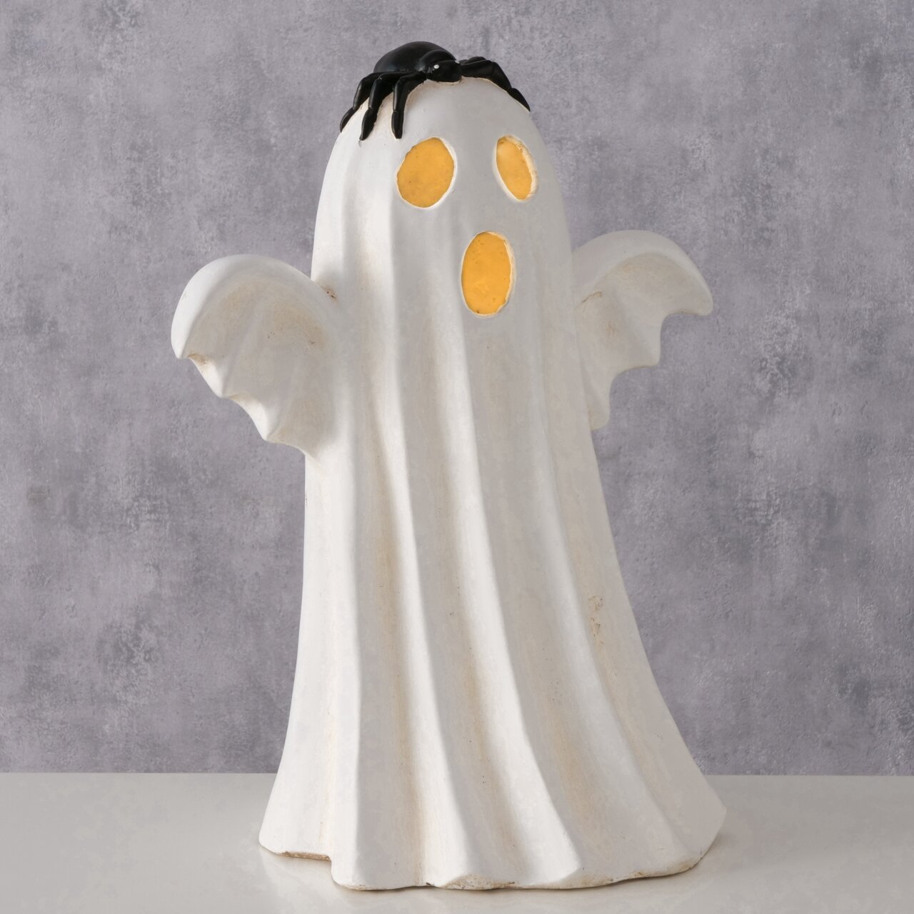 Weiße Halloween-Geisterfigur mit ausgebreiteten Armen, gelb leuchtenden Augen- und Mundöffnungen und schwarzer Spinne auf dem Kopf, frontal aus leicht erhöhter Perspektive vor grauem Hintergrund