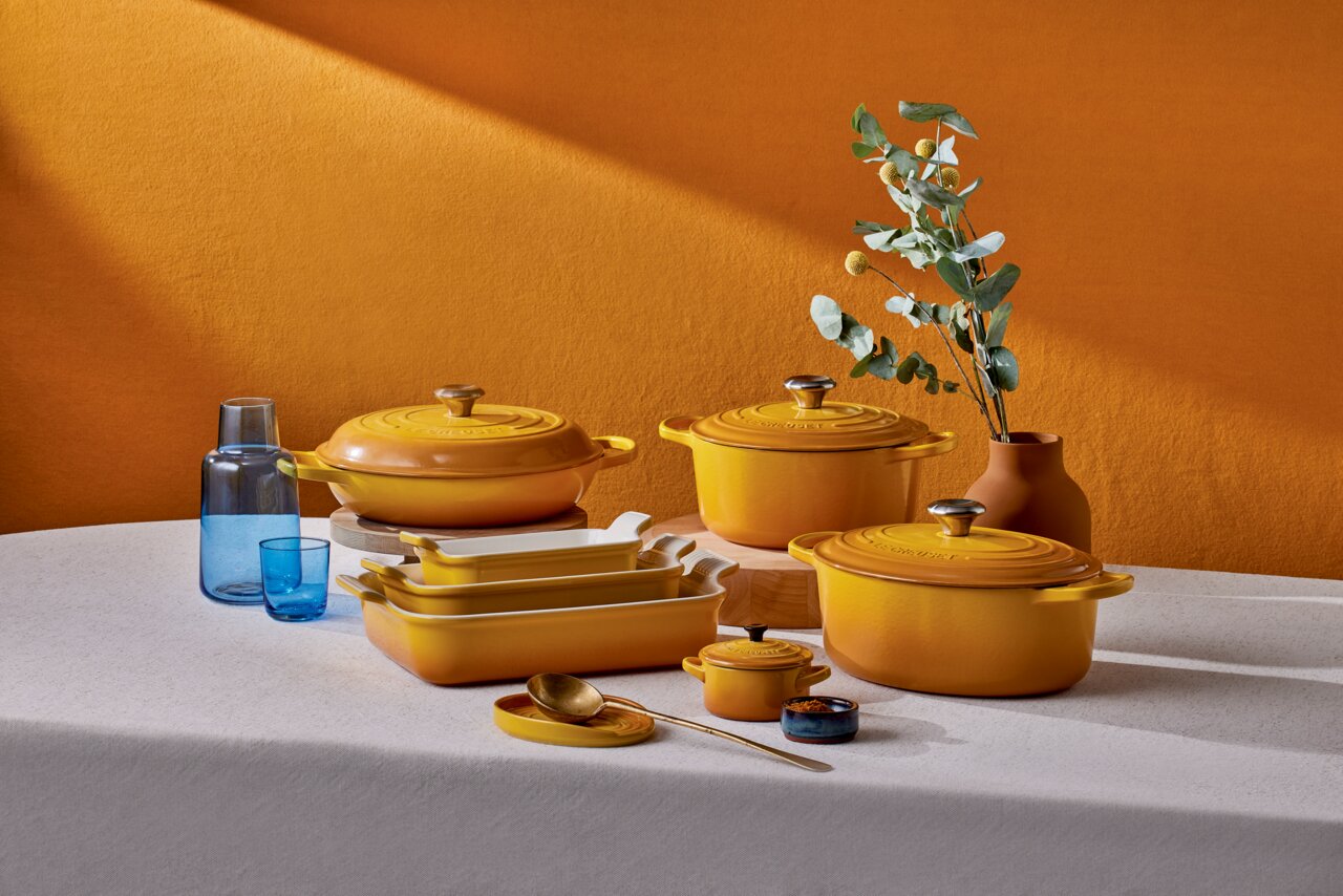 Ein Set aus gelben Brätern und Auflaufformen auf einem Tisch, umgeben von blauen Gläsern und einer Vase mit grünen Zweigen, aus einer seitlichen Perspektive fotografiert.
