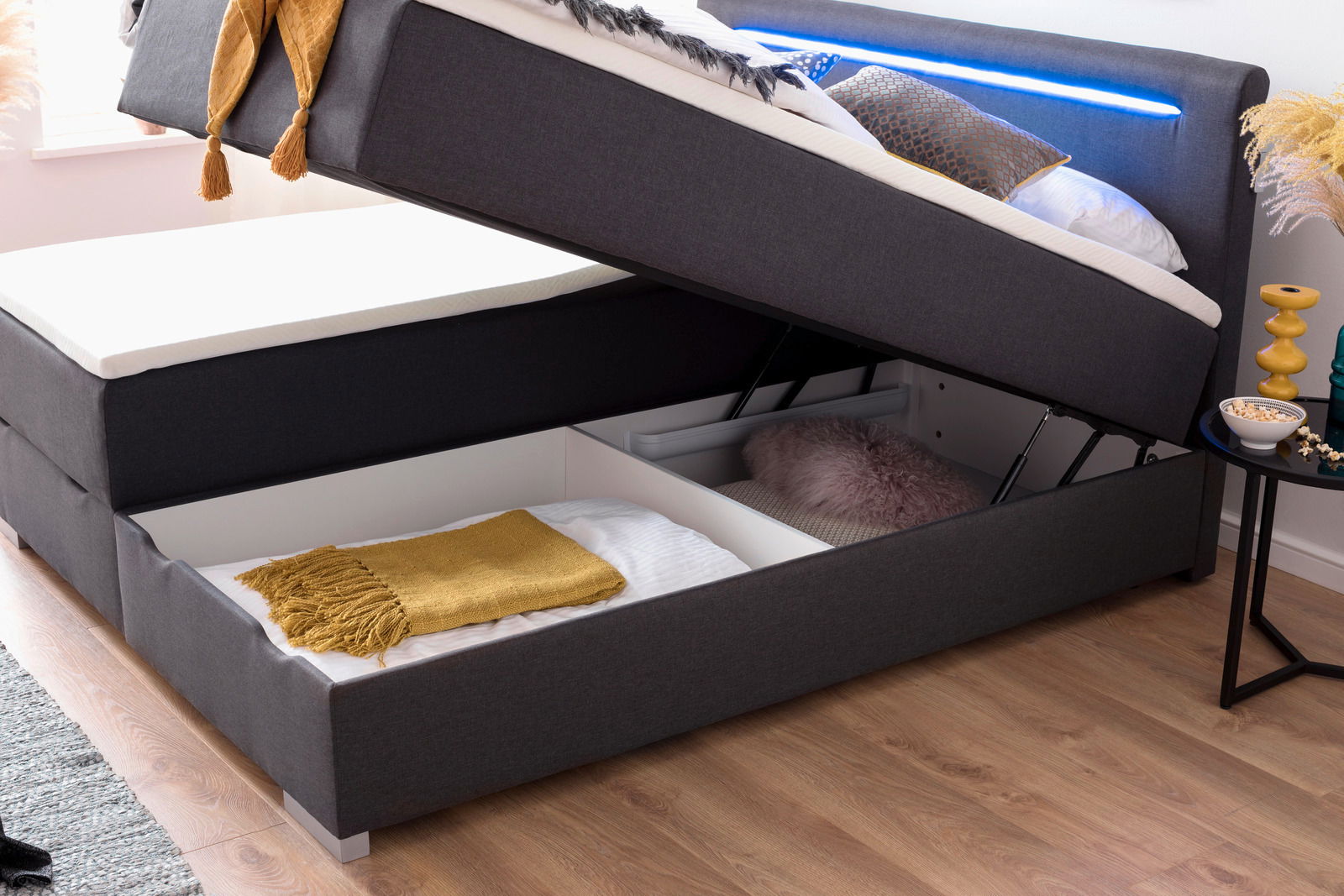 Boxbett mit hochgeklappter Matratze, darunter Stauraum mit Decken und Kissen, seitliche Perspektive