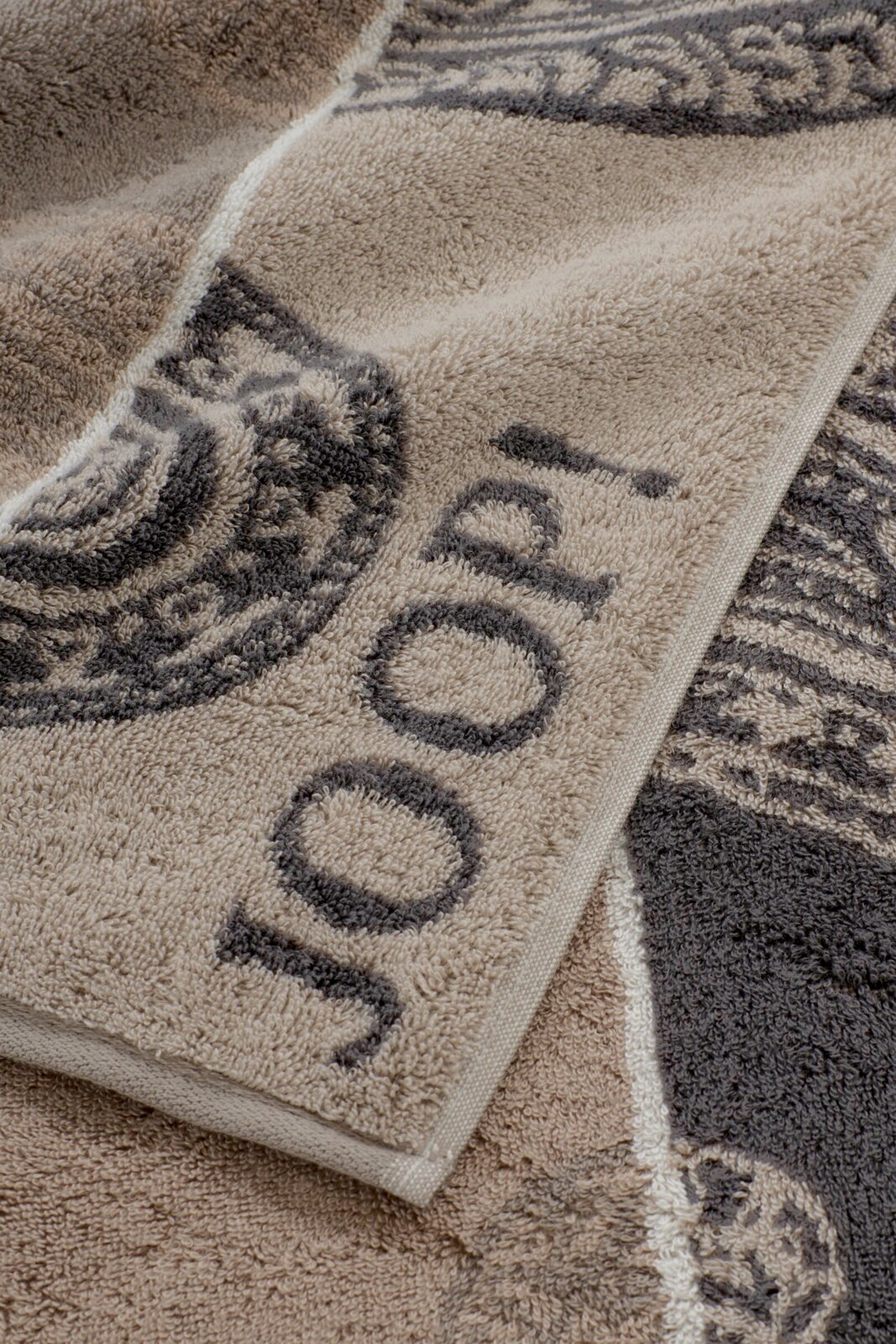 Nahaufnahme eines weichen Frottee-Handtuchs in Beige und Grau mit gewebtem Muster und Schriftzug, perspektivisch schräg von oben fotografiert