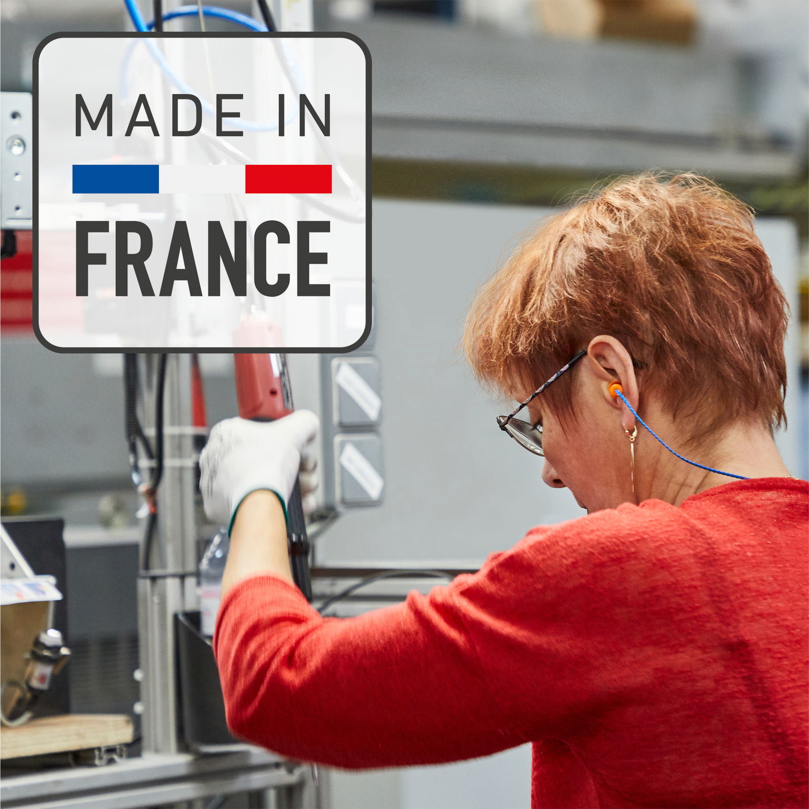 Frau in rotem Pullover arbeitet in einer Fabrik, mit einem 'Made in France' Schild im Vordergrund, Seitenansicht.