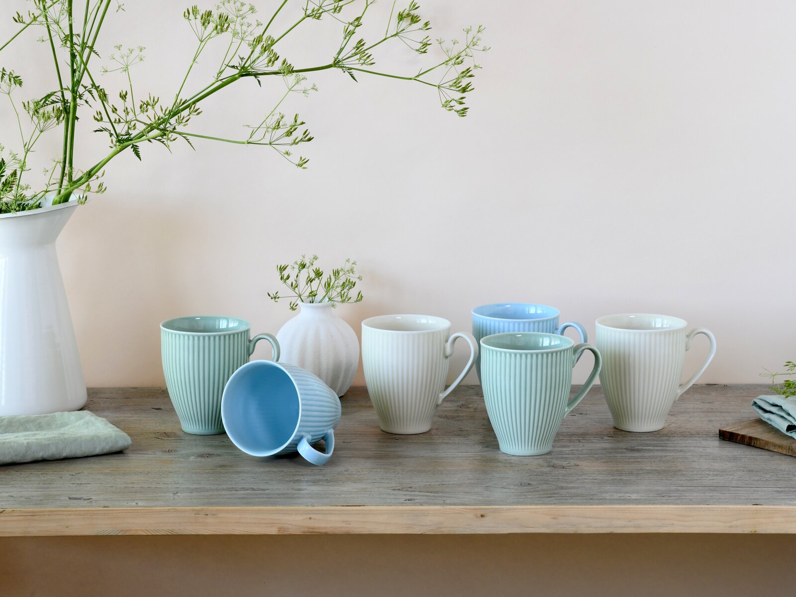 Pastellfarbene, geriffelte Keramik-Kaffeebecher mit Henkeln in Mint, Blau und Creme auf rustikalem Holztisch, ein Becher liegt seitlich; Frontansicht auf Augenhöhe.