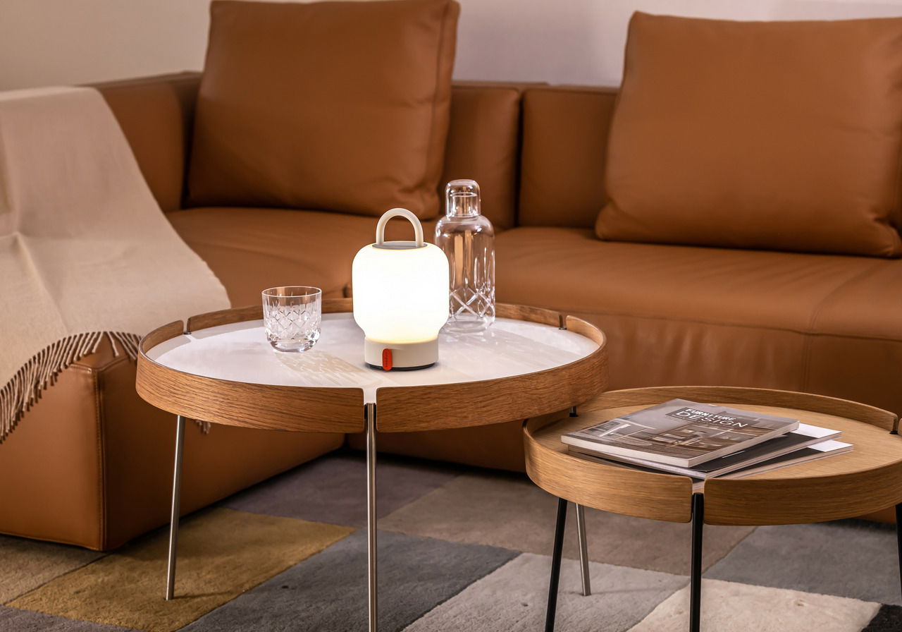 Moderne Tischlampe in einem gemütlichen Wohnzimmer, auf einem runden Holztisch platziert, mit einem Ledersofa im Hintergrund. Perspektive von vorne.