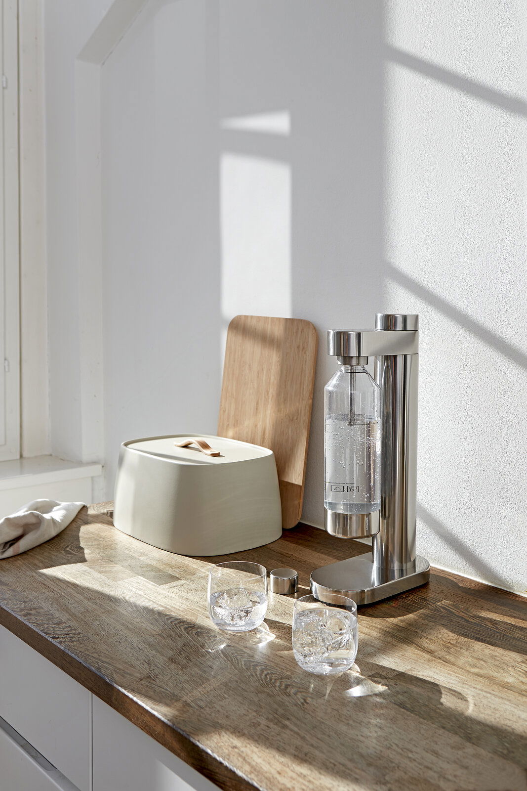 Stelton Brus Wassersprudler inkl. Wasserflasche - steel Brus Wassersprudler aus Edelstahl auf einer Küchenarbeitsplatte, seitliche Perspektive, mit zwei Gläsern Wasser und einem Brotkasten im Hintergrund.
