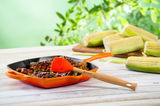 Grillpfanne in Ofenrot mit gegrilltem Fleisch und Silikonpinsel, seitliche Perspektive, im Hintergrund Maiskolben und grüne Blätter