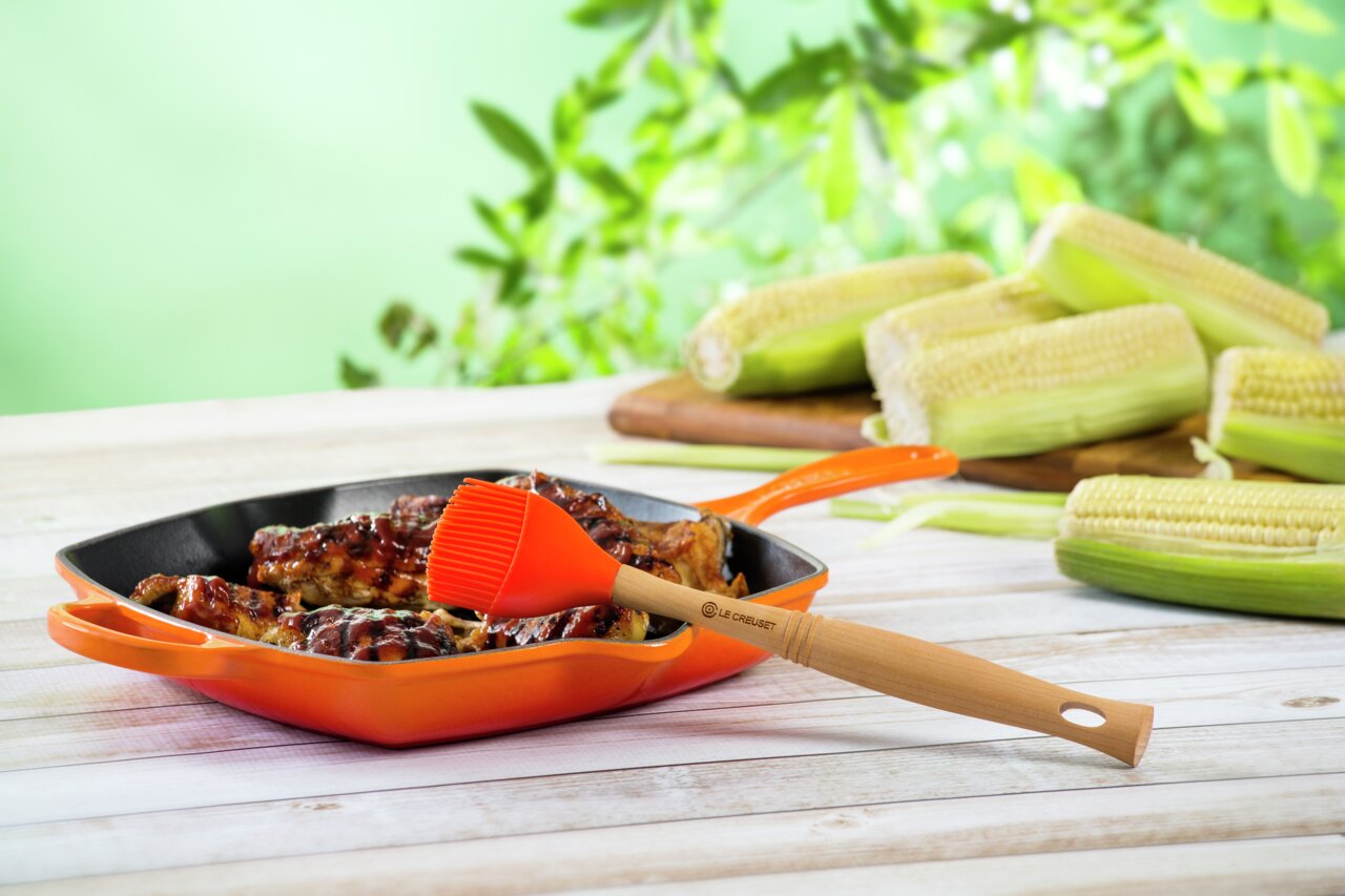 Grillpfanne in Ofenrot mit gegrilltem Fleisch und Silikonpinsel, seitliche Perspektive, im Hintergrund Maiskolben und grüne Blätter
