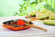Grillpfanne in Ofenrot mit gegrilltem Fleisch und Silikonpinsel, seitliche Perspektive, im Hintergrund Maiskolben und grüne Blätter