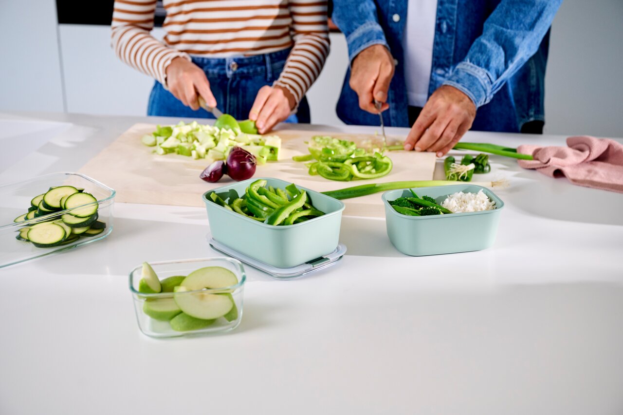 Twee mensen snijden groenten in een moderne keuken, terwijl er groene bewaardozen op het aanrecht staan. Vooraanzicht.