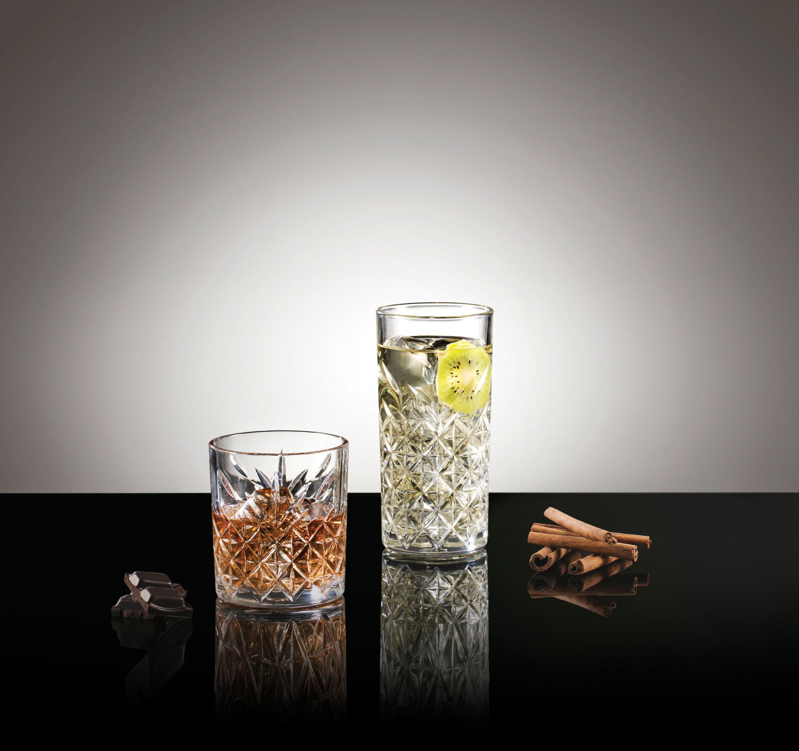Zwei Gläser aus dem Glasset Timeless 8-tlg. Set, eines niedrig und eines hoch, auf einer reflektierenden Oberfläche mit Zimtstangen und Schokolade, seitliche Perspektive.