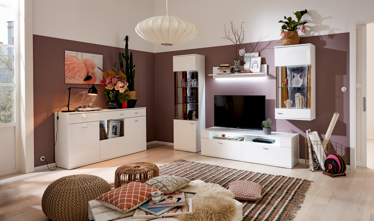 Set one by Musterring Vitrine re. TACOMA Moderne Wohnzimmer-Einrichtung mit weißer Vitrine rechts, dekoriert mit Gläsern, in einer gemütlichen Ecke mit Teppich und Kissen, aus frontaler Perspektive.