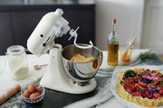Küchenmaschine in Cremefarbe mit Teighaken, seitliche Perspektive, auf einer Arbeitsfläche mit Zutaten wie Mehl, Eiern und einer Gemüsetarte.