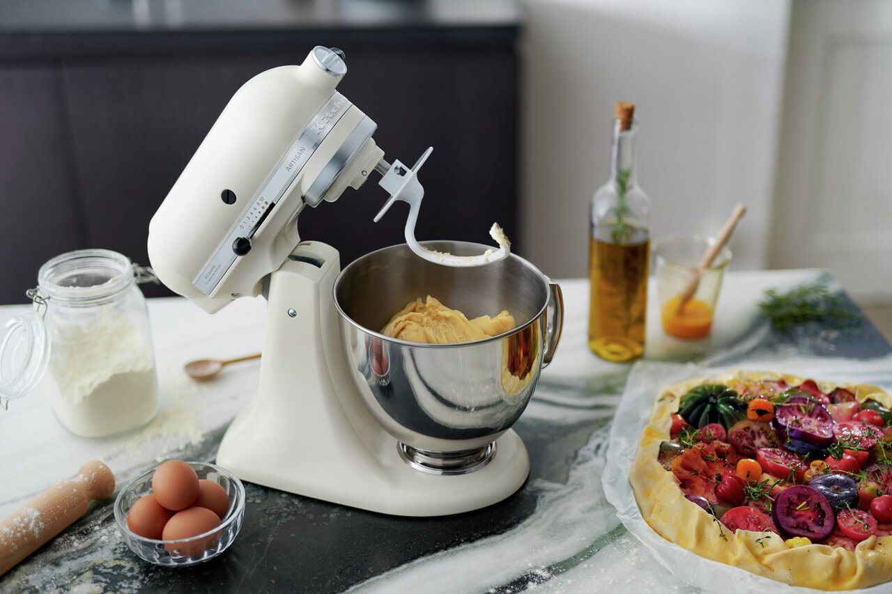 Küchenmaschine in Cremefarbe mit Teighaken, seitliche Perspektive, auf einer Arbeitsfläche mit Zutaten wie Mehl, Eiern und einer Gemüsetarte.