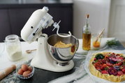 Küchenmaschine in Cremefarbe mit Teighaken, seitliche Perspektive, auf einer Arbeitsfläche mit Zutaten wie Mehl, Eiern und einer Gemüsetarte.
