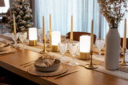 Festlich gedeckter Esstisch mit goldfarbenen Kerzenhaltern und Stabkerzen, warm leuchtenden Tischlampen, strukturierten Weingläsern, beigen Platzsets und weißer Vase; Perspektive: seitliche, leicht schräge Nahaufnahme entlang der Tischkante.