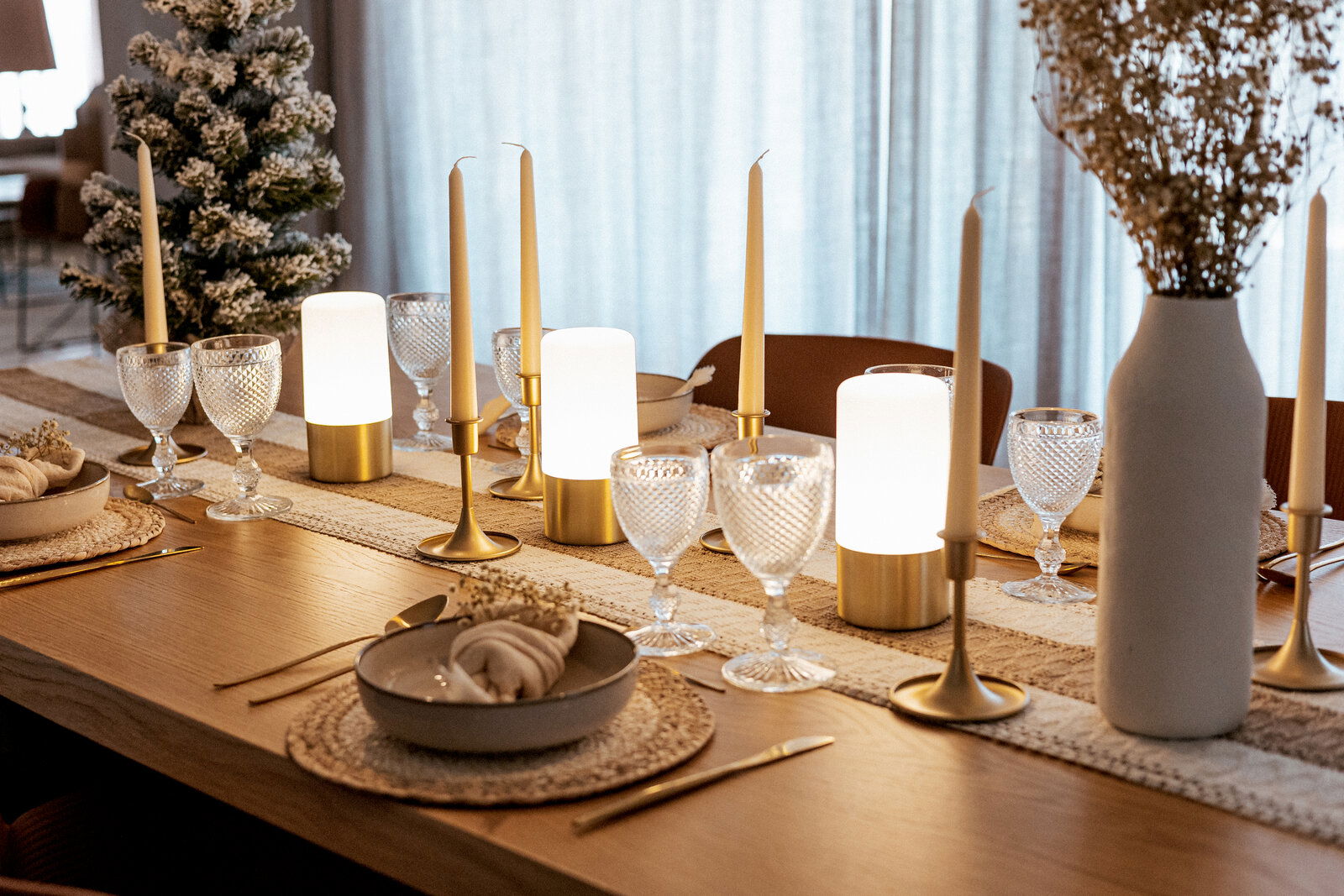 Festlich gedeckter Esstisch mit goldfarbenen Kerzenhaltern und Stabkerzen, warm leuchtenden Tischlampen, strukturierten Weingläsern, beigen Platzsets und weißer Vase; Perspektive: seitliche, leicht schräge Nahaufnahme entlang der Tischkante.