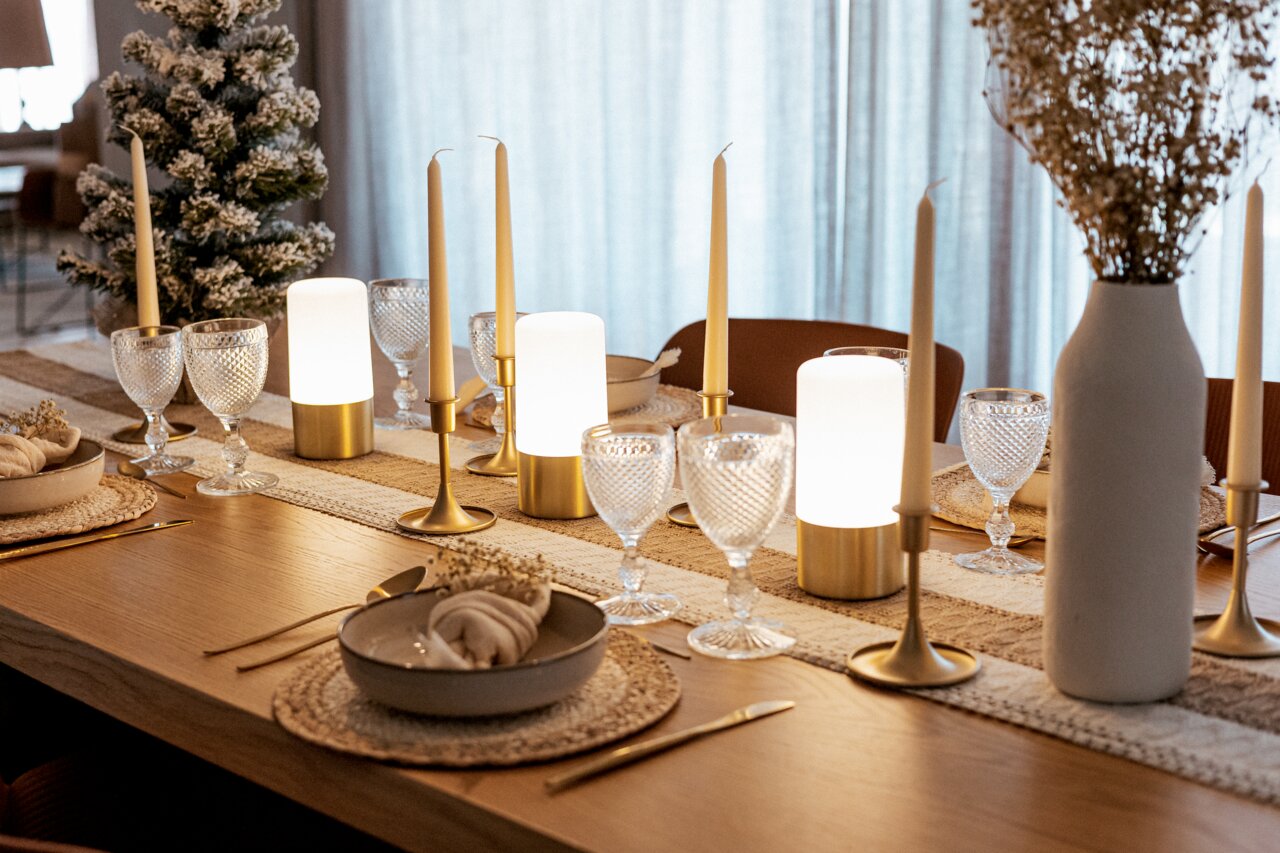 Een feestelijk gedekte eettafel met goudkleurige kandelaars en staafkaarsen, warm schijnende tafellampen, gestructureerde wijnglazen, beige placemats en een witte vaas perspectief: zijdelingse, licht schuine close-up langs de rand van de tafel.