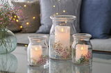 Drei Glasvasen mit LED-Kerzen und bunten Trockenblumen, auf einem Tisch in gemütlicher Wohnzimmeratmosphäre, von vorne betrachtet.