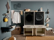 Frontalansicht eines modernen Hauswirtschaftsraums mit schwarzer Frontlader-Waschmaschine und Trockner in hellem Schrank, daneben Wäscheleinen, Körbe und Waschutensilien vor grauer Wand.