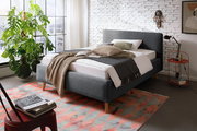 Modernes Polsterbett in Grau mit Holzfüßen, dekoriert mit Kissen und Decken, in einem stilvoll eingerichteten Raum mit Backsteinwand und großem Fenster, seitliche Perspektive.