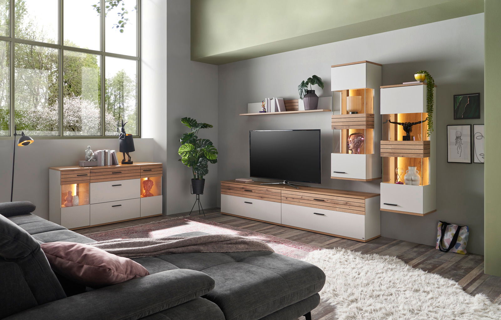 Set one by Musterring Sideboard SET ONE QUINCY Wohnzimmer mit moderner Möbelkombination, bestehend aus Sideboard, TV-Lowboard und Hängeschränken in Holz- und Weißtönen, mit dekorativer Beleuchtung und Pflanzen, seitliche Perspektive.