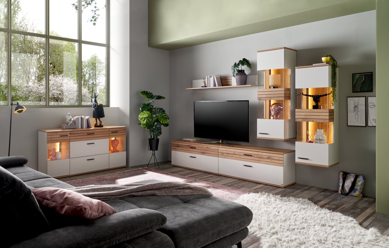 Wohnzimmer mit moderner Möbelkombination, bestehend aus Sideboard, TV-Lowboard und Hängeschränken in Holz- und Weißtönen, mit dekorativer Beleuchtung und Pflanzen, seitliche Perspektive.
