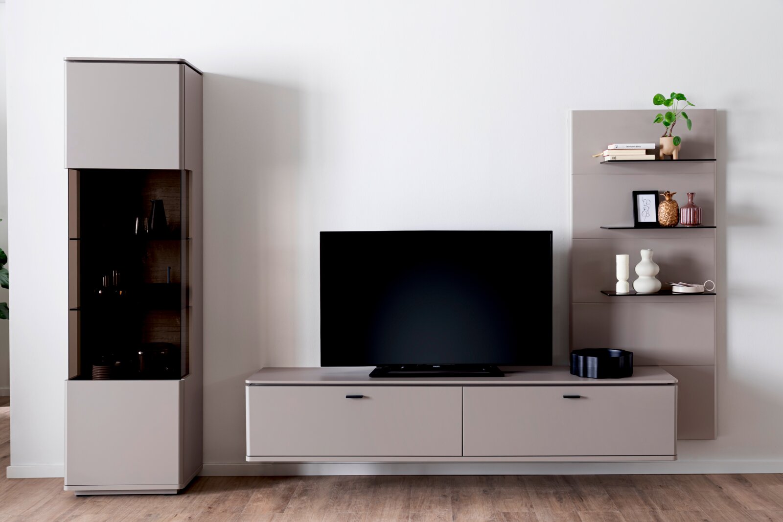 Modernes TV-Wohnwand-Set in Hellgrau mit Lowboard, Vitrine mit Glasfront und Wandregal mit Deko, frontale Perspektive auf die Möbel vor weißer Wand