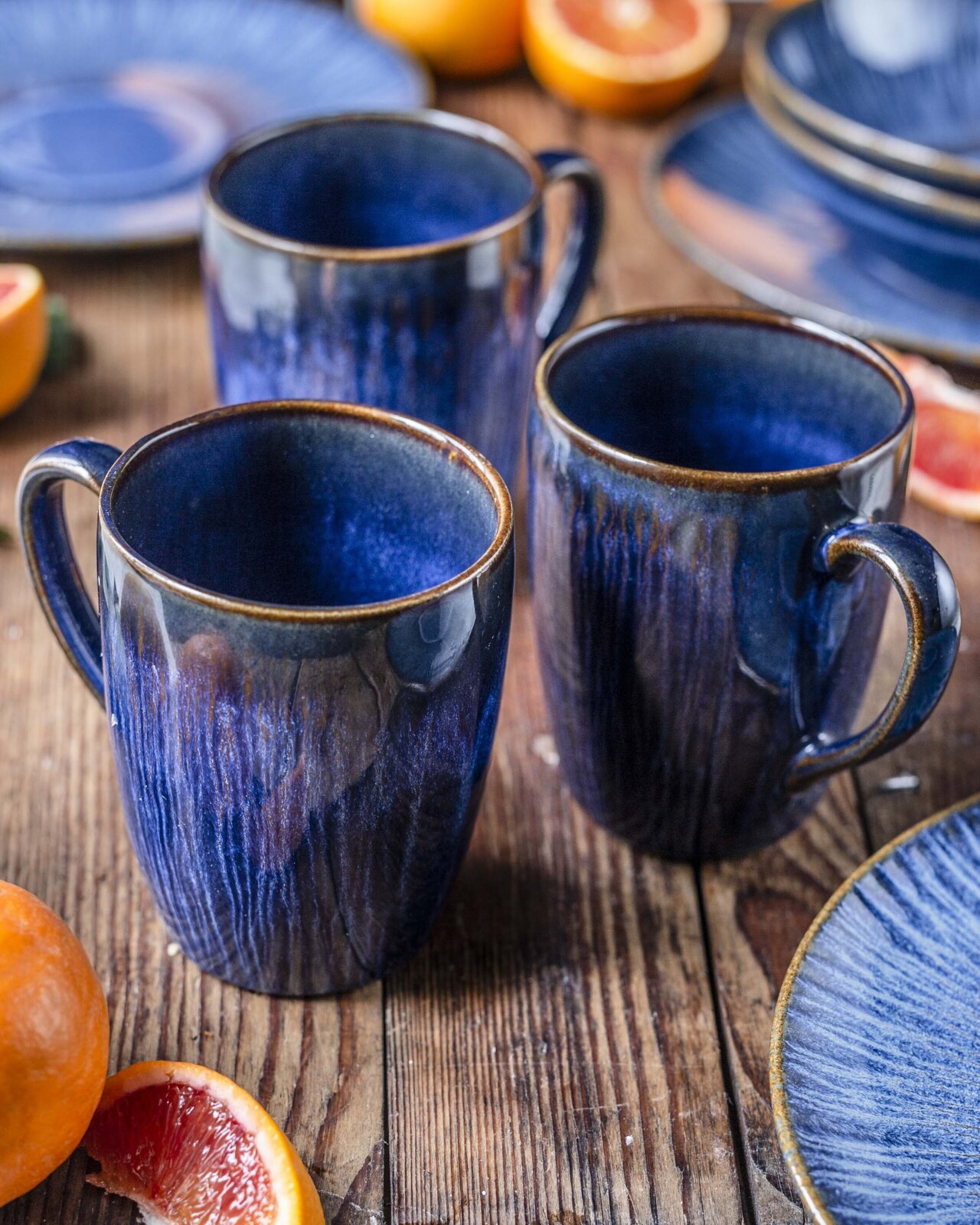 Drei dunkelblaue, glänzende Keramik-Kaffeebecher mit Henkel auf rustikalem Holztisch, umgeben von passenden Tellern und halbierten Zitrusfrüchten; Perspektive: leicht von oben in schräger Frontansicht.