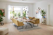 Moderner Esstisch mit Holzplatte und schwarzen Metallbeinen, umgeben von gelben Polsterbänken in einem hellen Wohnzimmer mit großen Fenstern, Blick von der Seite.